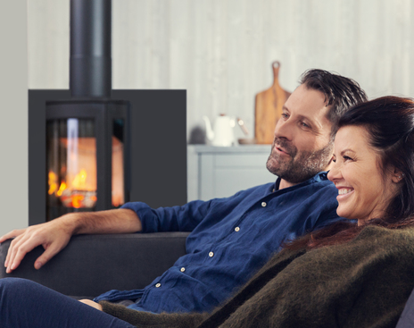 Un couple souriant est détendu sur un canapé avec une cheminée moderne allumée en arrière-plan, évoquant une scène de confort domestique pouvant se trouver en Vendée.