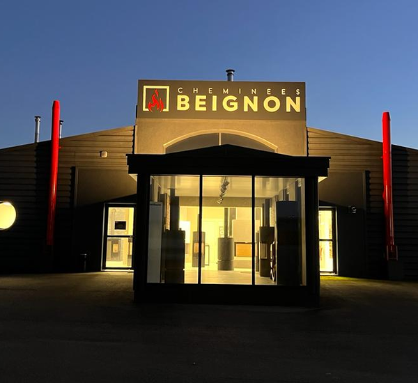 La façade du magasin "Cheminées Beignon" est éclairée par le soleil couchant, avec une enseigne lumineuse sur le toit et deux cheminées décoratives rouges de chaque côté, évoquant
